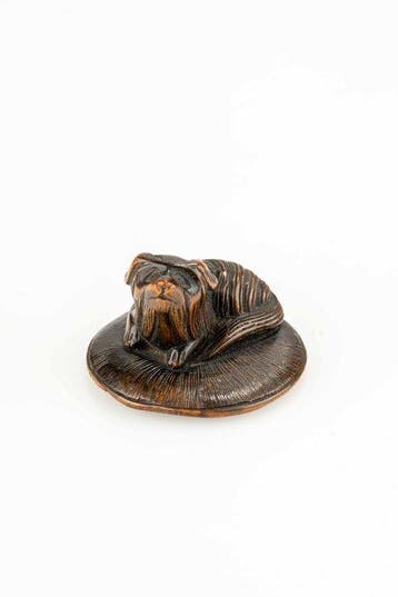 Een houten netsuke van een Pekinees pup die op een strohoed